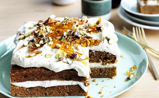 A square carrot cake, topped with cream-cheese icing and pistachios, on a eggshell-blue plate.