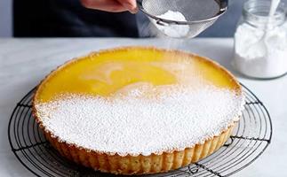 A whole lemon tart on a black wire rack being dusted with icing sugar.