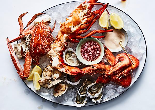 Over the top shot of a large white oval platter laden with seafood: a whole crab, a whole lobster, cooked prawns, clams, and shucked oysters. 