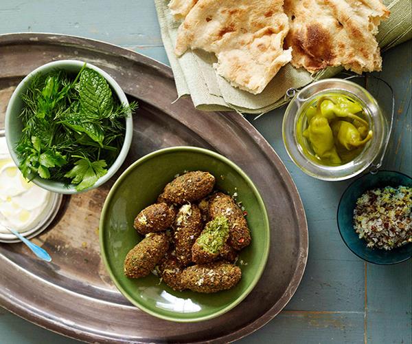 Bird's eye view of a feast, with flatbread in the top-right corner, an open jar of pickled chillies, a green bowl holding fried, oval-shaped falafel, and a white bowl holding a bowl of green herbs.