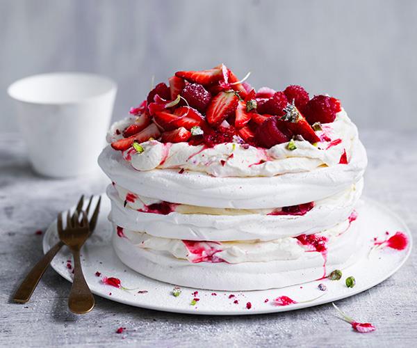 Three-tiered pavlova layered with berry jam, topped with strawberries and raspberries, sitting on a white plate with two brass forks on the side.
