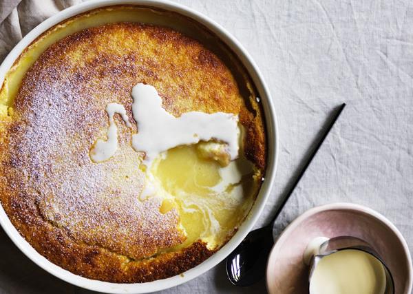 Over-the-top shot of a round white dish with a baked lemon pudding, with a spoonful taken out of the crust to reveal the molten lemon curd inside.