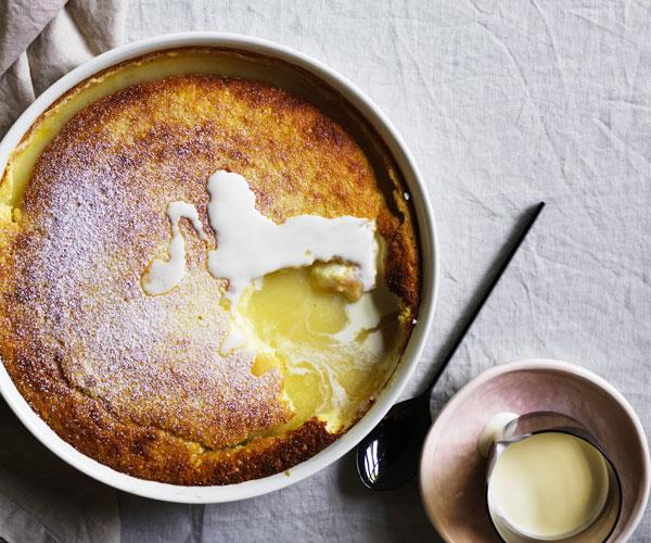 Over-the-top shot of a round white dish with a baked lemon pudding, with a spoonful taken out of the crust to reveal the molten lemon curd inside.