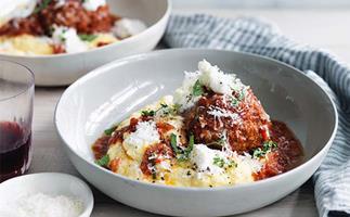 A wide grey bowl holding a pile of creamy polenta, topped with a large meatball, tomato sauce, chopped parsley and shaved parmesan.