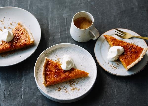 Three white round plates, each holding a triangular slice of golden-baked pumpkin pie, dusted with cinnamon, and served with a dollop of cream.
