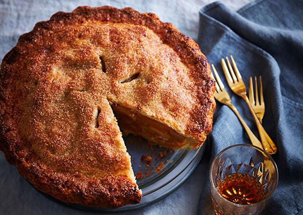 A round, crusty apple pie with a slice taken out of it, sitting on a light-blue linen tablecloth, with golden forks and a tumbler of amber-hued liquid to the side