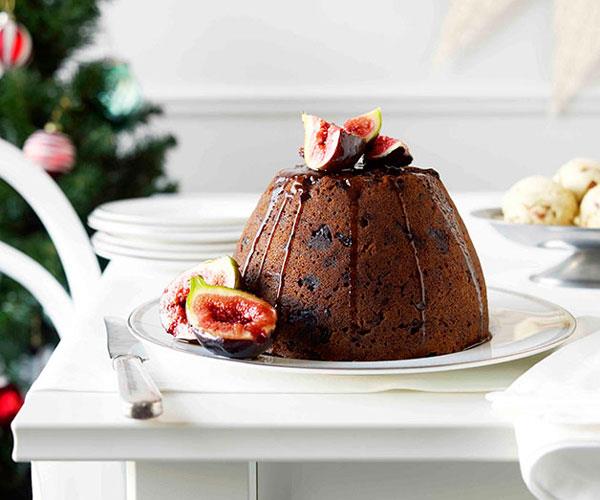 A boiled Christmas pudding, studded with dried fruit, topped with sliced figs and drizzled with a dark-brown port syrup, on a white plate, on a white table.