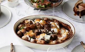 Photo of white baking dish filled with creamy potato bake surrounded by other plates and a glass of red wine