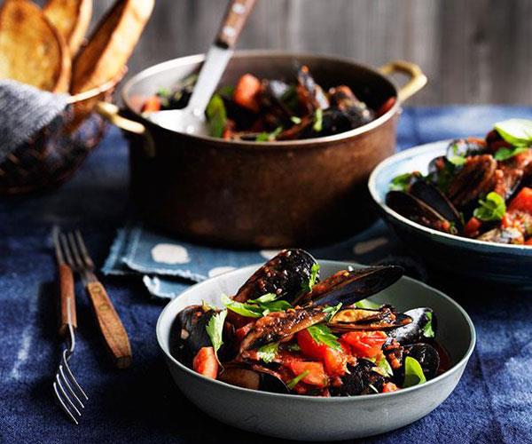 In the foreground, a grey dish holding a pile of steamed mussels with chopped tomatoes and basil, on a dark blue tablecloth. In the background, a copper pot holding more of said mussels.
