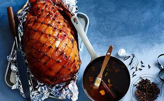 On a gradient blue background, there's a leg of ham in a roasting tray, the fat scored in a diamond pattern, studded with cloves, and brushed with a sticky glaze. A pot of glaze and a bowl of cloves sit on the side. 