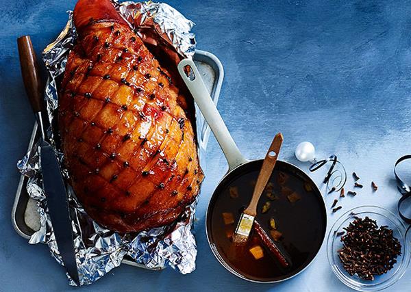 On a gradient blue background, there's a leg of ham in a roasting tray, the fat scored in a diamond pattern, studded with cloves, and brushed with a sticky glaze. A pot of glaze and a bowl of cloves sit on the side. 