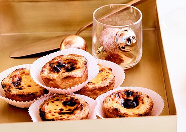 Golden square tin holding hand-sized custard tarts with burnished tops, each in their own white paper casing. There's also a glass holding a rose-pink speckled bauble, which is kind of weird, but we'll take it.