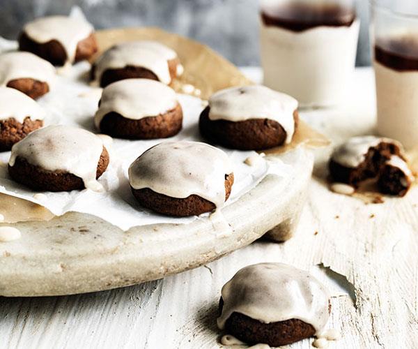 Brown, round pfeffernüsse biscuits, topped with icing, scattered on a raised white platter and white wooden table.