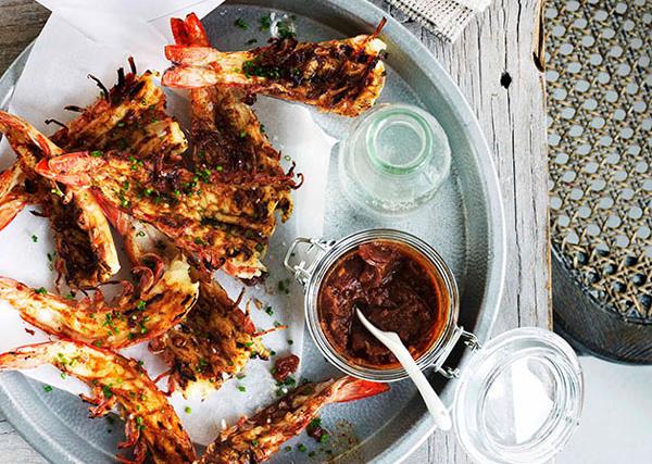 Over-the-top shot of a round dish holding butterflied and barbecued prawns, with satisfying grill marks, and scattered with green herbs, with a pot of dark and chunky spiced tomato sauce.