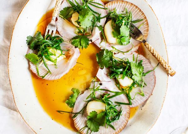 Five scallops in a half-shell topped with herbs and a soy dressing, on an off-white plate, on a white marble background. 