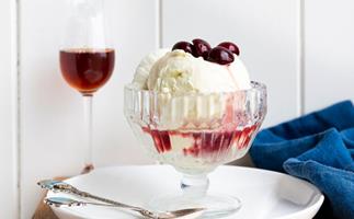 photo of wooden table and white wall with champagne flute filled with red liquid and a glass footed ice cream bowl with white ice cream, red liquid and topped with dark cherries in foreground