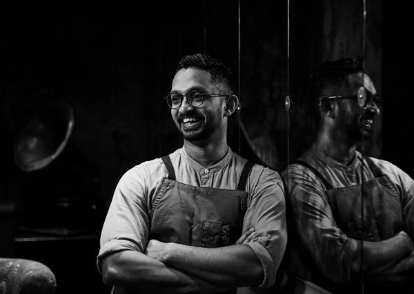 Black and white portrait of chef Rishi Naleendra leaning against a reflective wall.