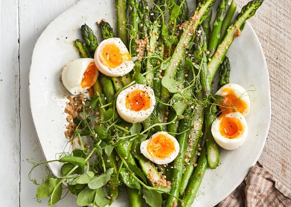 White plates with asparagus spears, halved soft-boiled eggs, and pea shoots, scattered with buckwheat.