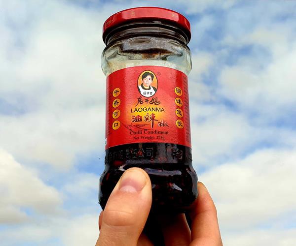 A jar of La Gan Ma chilli condiment against a blue sky with clouds.
