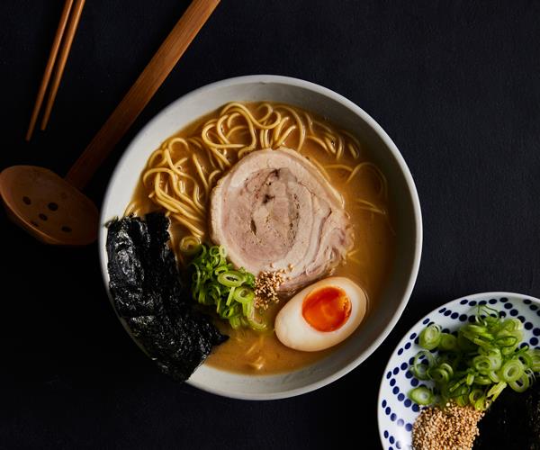 Overhead shot of a bowl of ramen topped with half a boiled egg, a slice of pork, nori and chopped spring onions, on a black background