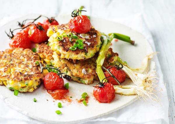A white plate with fried corn fritters and roasted cherry tomatoes, garnished with chopped and whole shallots.