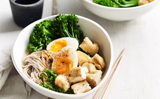 A white bowl with soba noodles, egg, tofy and broccolini, with a pair of wooden chopsticks on the side