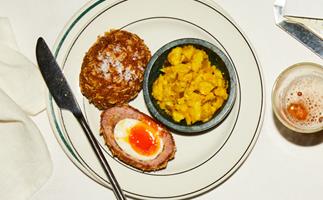 A halved Scotch egg, with a runny yolk centre, with a bowl of yellow-coloured cauliflower pickle, on a white round plate with a thin green border, on a white table. A stainless steel butter knife lies to the left, a glass of beer on the right.