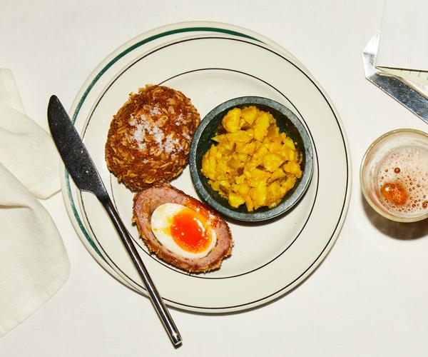 A halved Scotch egg, with a runny yolk centre, with a bowl of yellow-coloured cauliflower pickle, on a white round plate with a thin green border, on a white table. A stainless steel butter knife lies to the left, a glass of beer on the right.