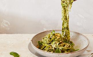 A raised fork dangling strands of long pasta coated in a green herb sauce, from a brown-white bowl. 