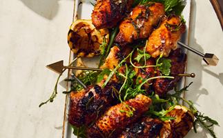 A rectangle plate holding skewers of charred, sticky chicken wings garnished with green herbs and a charred lemon half.