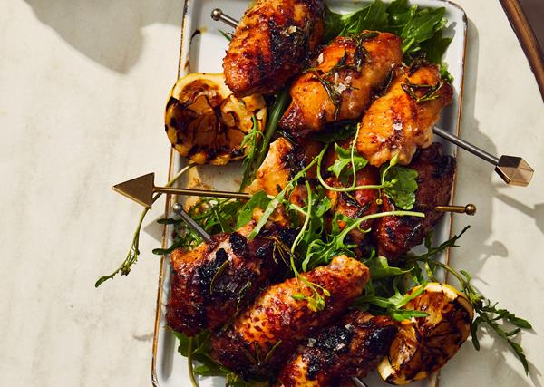 Skewers of barbecued chicken wings and lemon halves on a white rectangle tray, on an off-white marble table
