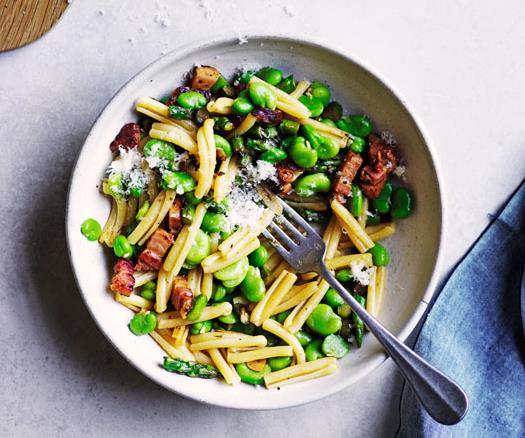 White bowl holding a mixture of short pasta, broad beans, asparagus and pancetta, with a steel fork inside, and a blue napkin to the side.
