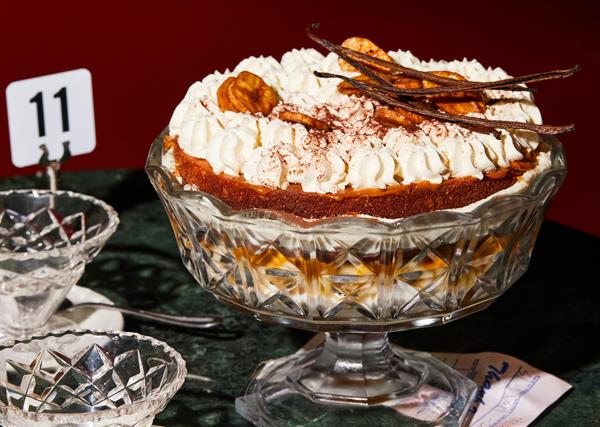 Ornamental glass bowl on a stand holding a trifle topped with whipped cream, banana chips and vanilla bean pods