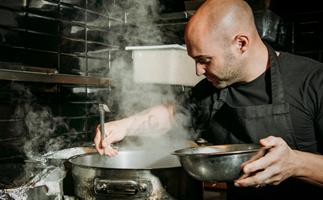 Mid-shot of a male chef in a black apron, stirring a large steel pot, with steam rising from it. 