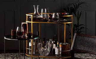 A two-level golden bar cart stocked with bottles of spirits, wine glasses and wine decanter. 
