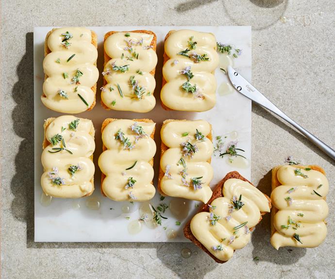 Over-the-top shot of white square plate holding eight small, rectangular shaped cakes topped with squiggles of light-yellow cream, and sprinkled with rosemary leaves and flowers