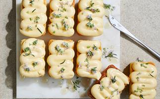 Over-the-top shot of white square plate holding eight small, rectangular shaped cakes topped with squiggles of light-yellow cream, and sprinkled with rosemary leaves and flowers