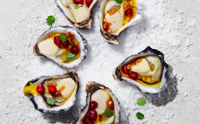 Over the top shot of six shucked oysters, arranged in a circle, on a bed of salt. Each oyster is topped with a pomegranate dressing and tiny green herb leaves.