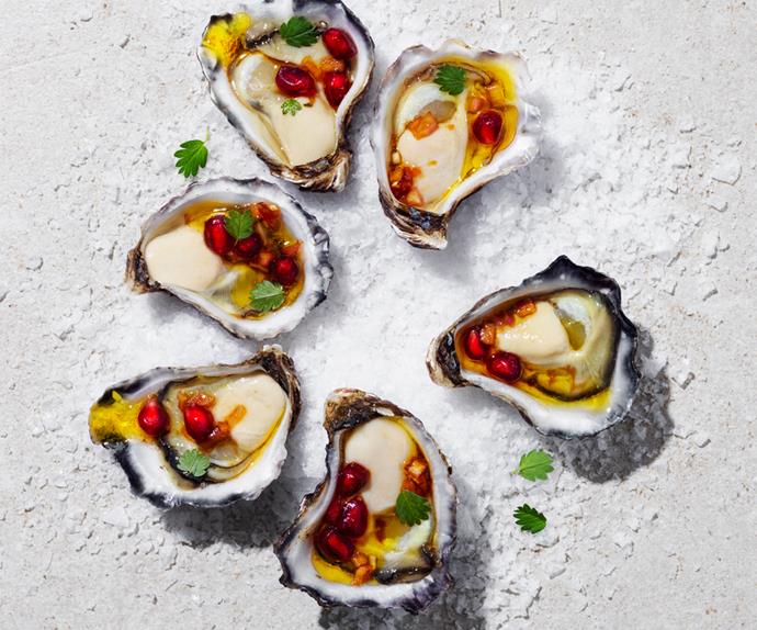 Over the top shot of six shucked oysters, arranged in a circle, on a bed of salt. Each oyster is topped with a pomegranate dressing and tiny green herb leaves.
