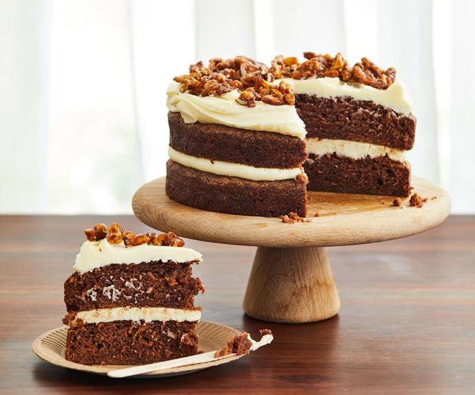 A two-layer carrot cake with cream cheese frosting, topped with walnuts, on a wooden cake stand. A thick triangle slice of carrot cake is in the foreground, on the left.