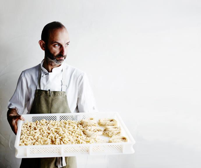 Chef Andreas Papadakis in a white shirt and olive-green apron holding a tray of freshly made pasta.
