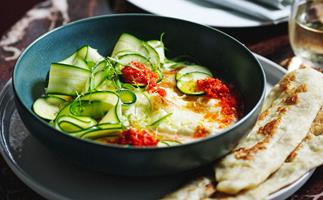 A dark green bowl with baked burrata, chilli sauce and slivers of zucchini, with some flatbread to the side.
