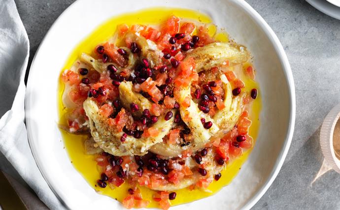Overhead shot of grilled eggplant slices, finely chopped tomato, pomegranate seeds and olive oil on a round white plate.