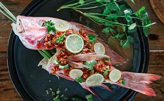 A whole steamed snapper on a black plate, garnished with sliced limes, crushed chillies and coriander sprigs.