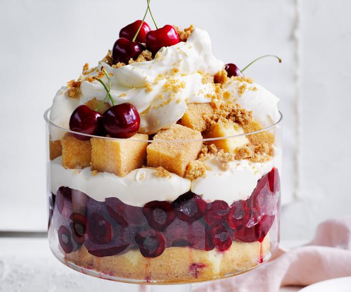 Large, raised glass bowl holding a trifle layered with sponge cake, halves cherries, cream and crumbs