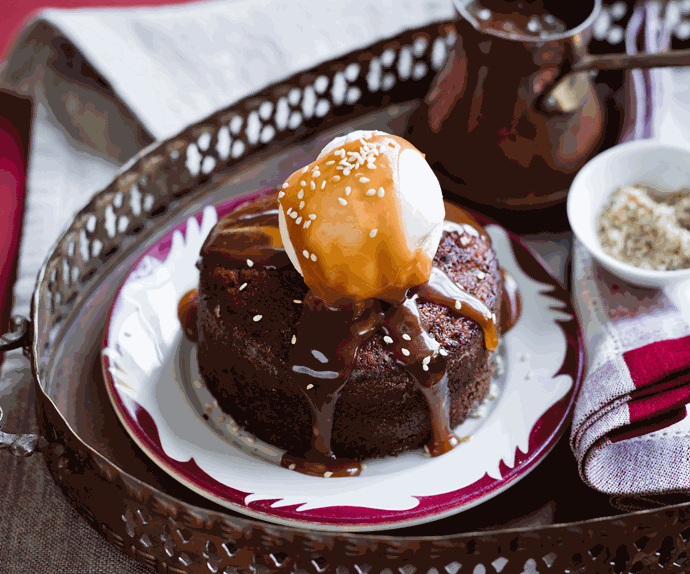A circular date pudding topped with a scoop of sesame ice-cream and drizzled with salted caramel. A pot of salted caramel featured behind, and a bowl of toasted sesame seeds.