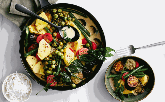Generous chunks of eggplant, pineapple, tomatoes, snake beans, and pea eggplants in coconut curry sauce in a black saucepan.
