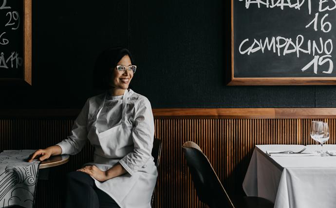 Portrait image of female chef in white apron sitting at restaurant table with blackboards with chalk writing in background