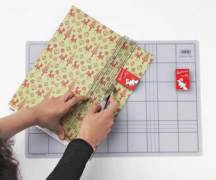 **Step Two:** Cut out a piece of fabric and cover some foam or cardboard with it – this will form the backdrop for your Advent calendar. Decorate with strips of ribbon and push pins. Empty out 12 matchboxes. Using a ruler and box cutter, cut different decorative wrapping paper.
