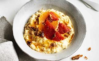 Rice pudding, topped with orange segments and crushed sesame brittle, in a grey bowl, with a grey linen napkin to the left.